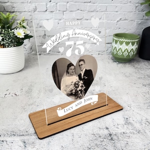 May include: A clear acrylic photo frame with a wooden base. The frame features a heart-shaped photo of a couple with the text "Happy Wedding Anniversary 75" and "Lucy and John" below the photo.