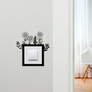 May include: A black square light switch cover with a white switch, adorned with a floral design. The decorative flowers and leaves are cut from black material, creating a contrasting aesthetic against the white wall.