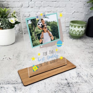 May include: A clear acrylic photo frame with a wooden stand. The frame features a photo of a father and son, with the text "For the best Daddy in the world" and stars and a rocket ship.