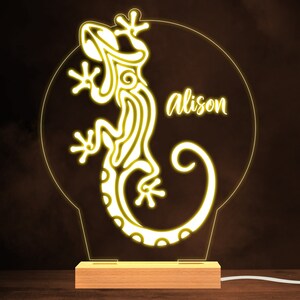 Può includere: Una luce al neon gialla con un design di geco stilizzato e il nome "alison" in un font corsivo. La luce è su una base di legno.