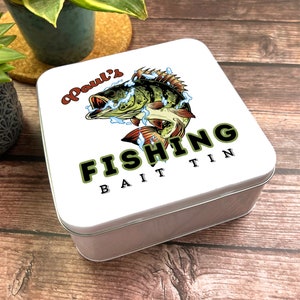 Può includere: Scatola di metallo bianca con un'illustrazione verde e marrone di un pesce che salta fuori dall'acqua. Il testo "Paul's Fishing Bait Tin" è stampato sulla scatola.