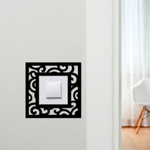 Peut inclure: Un cache-interrupteur noir de forme carrée avec un motif ornemental. Il entoure un interrupteur blanc sur un mur blanc. Le design présente des motifs tourbillonnants. L'image montre un aperçu d'une pièce.