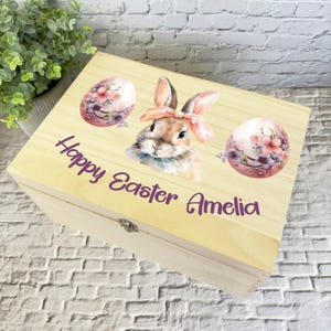 Puede incluir: Una caja de madera con tapa con bisagras, decorada con una ilustración de acuarela de un conejo, flores y huevos de Pascua. La caja está personalizada con el texto "Feliz Pascua Amelia".