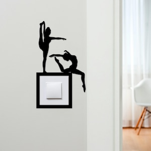 May include: Black silhouette wall art of two gymnasts in graceful poses, one standing with a leg extended and the other seated. The art is mounted on a light switch with a black frame, adding a decorative touch to the wall.