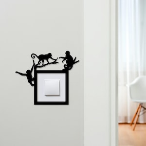 Puede incluir: Arte mural de metal negro con tres siluetas de monos en ramas, rodeando un interruptor de luz. Los monos están en poses juguetonas, creando un acento decorativo y caprichoso. El interruptor es blanco y está enmarcado en negro.