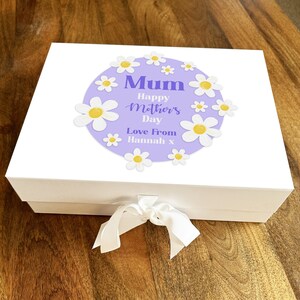 Può includere: Scatola regalo bianca con un cerchio viola e margherite bianche. Il cerchio contiene il testo "Mum Happy Mother's Day Love From Hannah x".