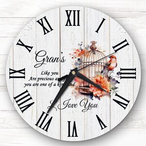 May include: A white wooden clock with Roman numerals and a black clock hand. The clock face has a watercolor design of a bird in a cage with flowers. The text "Gran's" is written in black cursive font. The text "Like you are precious and you are one of a kind" is written in black font. The text "Love You" is written in black cursive font.