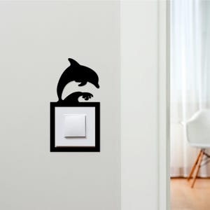 May include: Black dolphin silhouette leaping over a wave, mounted on a square frame. The frame surrounds a white light switch. The design is affixed to a white wall, adding a decorative touch to a functional item.