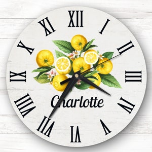 May include: White round clock with Roman numeral markings and black hands. The clock face features a lemon branch with yellow lemons and green leaves. The name "Charlotte" is written in black cursive below the lemons.