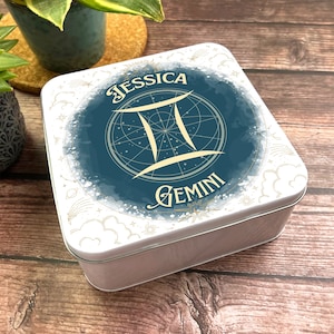 Könnte beinhalten: Eine weiße quadratische Dose mit einem blauen und goldenen Design. Das Design zeigt das Sternzeichen Zwillinge, das Wort "Zwillinge" und den Namen "Jessica".