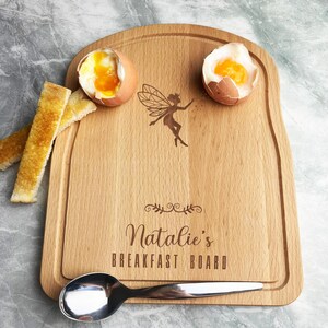 Könnte beinhalten: Ein Frühstücksbrett aus Holz mit einem Feen-Design und dem Text "Natalie's BREAKFAST BOARD". Das Brett hat eine vertiefte Kante und enthält zwei weichgekochte Eier, Toast und einen Löffel. Die Eier haben leuchtend gelbe Eigelb.