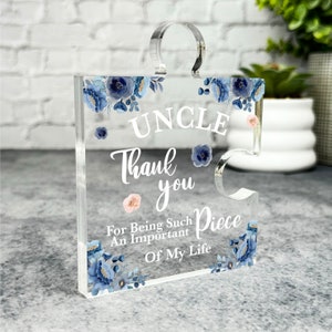 Puede incluir: Una pieza de rompecabezas de acrílico transparente con un diseño floral en tonos azules y rosas. El texto "Tío Gracias por ser una pieza tan importante de mi vida" está impreso en la pieza.