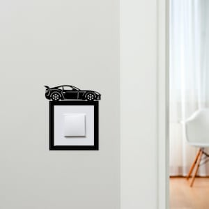 May include: Black car-shaped light switch cover with a square frame and a white switch. The car has a spoiler and detailed wheels. The cover is mounted on a white wall.