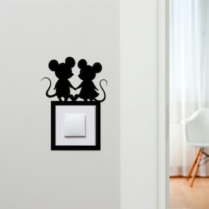 May include: Black silhouette of two mice holding hands, perched above a light switch. The mice are detailed with large ears and curly tails. The light switch is set in a black frame, creating a decorative accent for a wall.