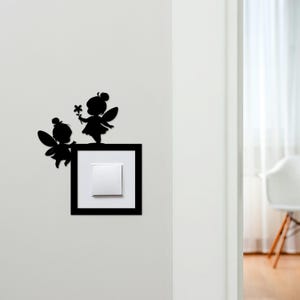 May include: Black silhouette fairy wall art decor. Two fairies with wings and flower accents are perched above a light switch. The light switch is framed in a black square, creating a whimsical accent for any room.
