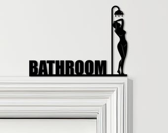 Könnte beinhalten: Schwarzes Metall-Badezimmerschild mit dem Wort "BATHROOM" und einer Silhouette einer Person unter einem Duschkopf. Das Schild ist an einem weißen Türrahmen befestigt.