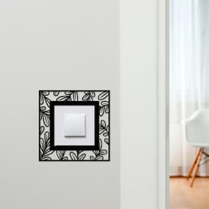 Può includere: Una copertura decorativa per interruttore della luce con un design floreale nero. La cornice quadrata circonda un interruttore bianco e un quadrato interno bianco. Il design è montato su una parete bianca.