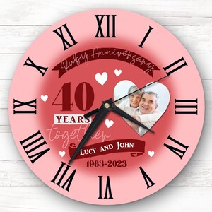 May include: Pink round clock with Roman numerals. The clock face has a red heart with a photo of a couple inside. The text on the clock reads "Ruby Anniversary 40 YEARS together LUCY AND JOHN 1983-2023".