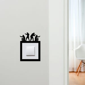 May include: A black metal light switch cover with a silhouette design of four figures pointing guns. The cover is rectangular with a white square switch in the center. The design is mounted on a white wall.