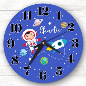 Op de afbeelding: Een ronde blauwe wandklok met zwarte cijfers en wijzers. De wijzerplaat toont een astronaut, planeten, een raket en de naam "Charlie" in witte letters. De klok heeft een ruimtethema.