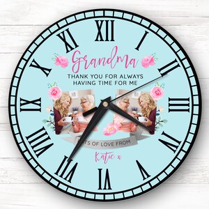 May include: A round wall clock with a light blue face, black Roman numerals, and black hands. The clock features the word "Grandma" in pink script, floral accents, and photos. The text reads "THANK YOU FOR ALWAYS HAVING TIME FOR ME."
