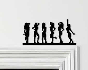 Könnte beinhalten: Eine schwarze Metall-Silhouette von sechs Figuren in verschiedenen Posen. Die Figuren stehen auf einer horizontalen schwarzen Basis, mit einem erhobenen Arm. Die Figuren tragen alle High Heels. Die Silhouette ist auf einer weißen Oberfläche montiert.