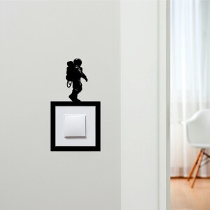 May include: A black silhouette of an astronaut walking on a black framed light switch. The light switch is white. The astronaut is wearing a helmet and a backpack. The wall is white.
