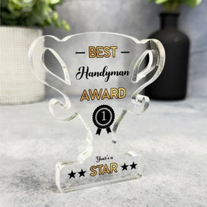 May include: A clear acrylic trophy with the text "Best Handyman Award" and a gold star on the base. The trophy has a black number 1 on a gold circle with a black ribbon.