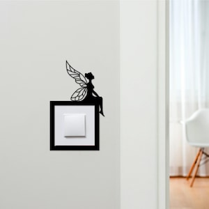 May include: A black metal fairy silhouette decorates a light switch. The fairy is perched on a black square frame surrounding a white light switch. The fairy has detailed wings and is sitting with her legs crossed.