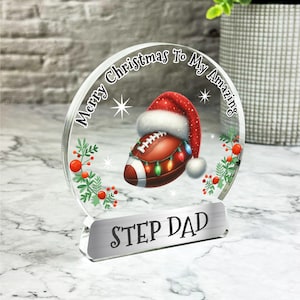 May include: A clear glass ornament with a red and white Santa hat and a football with Christmas lights. The ornament has the text "Merry Christmas To My Amazing Step Dad" on it.