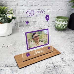 May include: A clear acrylic photo frame with a purple border and a wooden base. The frame has a photo of a man smiling and the text "50 years" with a list of time measurements in months, weeks, days, hours, minutes, and seconds. The text "James" is below the photo.