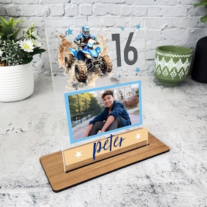 Puede incluir: Un marco de fotos de acrílico transparente con una base de madera. El marco presenta una ilustración de acuarela de una persona conduciendo un ATV, el número 16 y estrellas azules. Una foto de una persona se encuentra en la parte inferior del marco. El nombre "peter" está escrito debajo de la foto.