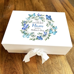 Puede incluir: Caja de regalo blanca con tapa, atada con una cinta blanca. La tapa presenta una corona de mariposas azules y vegetación, con el texto "For a wonderful Mum Happy Mother's Day".