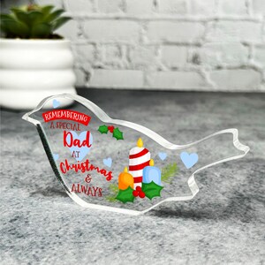 Może przedstawiać: Transparentna ozdoba ptaka z akrylu z napisem "Remembering a special Dad at Christmas & Always" w czerwonych i niebieskich literach. Ozdoba przedstawia czerwoną i białą świecę w paski, niebieską świecę i liście ostrokrzewu.