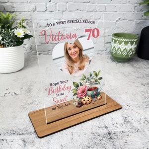 Może przedstawiać: Przezroczysta akrylowa ramka na zdjęcia z drewnianą podstawą. Ramka zawiera zdjęcie kobiety, imię "Victoria" i liczbę "70". Ilustracje kwiatowe i filiżanki do herbaty towarzyszą tekstowi "Hope Your Birthday is as Sweet as You Are."