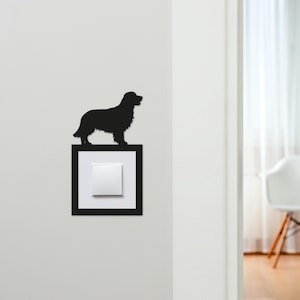 May include: A black silhouette of a dog, likely a Golden Retriever, perched atop a light switch. The light switch is set in a black frame against a white wall. The dog's outline is detailed, and the overall design is minimalist.