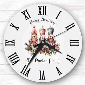 Könnte beinhalten: Weiße runde Uhr mit römischen Ziffern für die Stunden. Das Zifferblatt der Uhr zeigt eine Aquarell-Illustration von drei Nussknacker-Figuren mit einem Kranz aus Stechpalme und Beeren. Der Text "Merry Christmas" und "The Parker family" ist auf dem Zifferblatt der Uhr gedruckt.