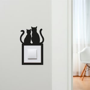 May include: Black cat silhouette wall decor, featuring two cats perched above a light switch. The cats are in a sitting position with curved tails. The light switch is white, set in a black frame, mounted on a white wall.