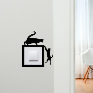 May include: A black light switch cover with a cat theme. The cover has a black frame, a white switch, and two black cat silhouettes. One cat is on top, and the other is climbing the frame. This design adds a playful element to any room.