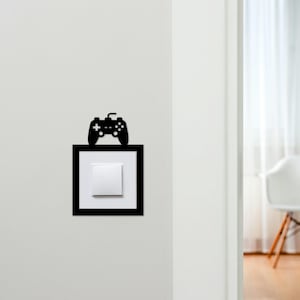 May include: A black decorative wall art piece featuring a video game controller silhouette above a light switch. The light switch is white, set within a black square frame. The wall is painted white.