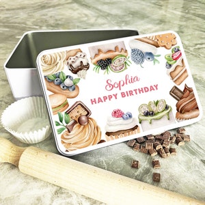 Puede incluir: Lata de metal blanca con un diseño acuarela colorido de varios cupcakes y pasteles. La lata tiene el texto "Sophia Feliz Cumpleaños".