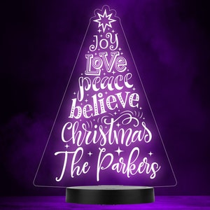 May include: White illuminated Christmas tree shaped acrylic sign with the words "Joy, Love, Peace, Believe, Christmas, The Parkers" on a black base.
