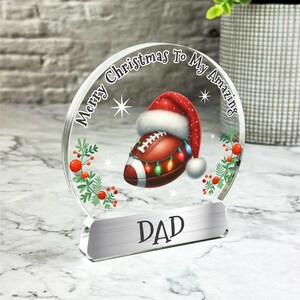 May include: A clear glass Christmas ornament with a red and white Santa hat on top of a football with Christmas lights. The ornament has the text "Merry Christmas To My Amazing Dad" around the edge.