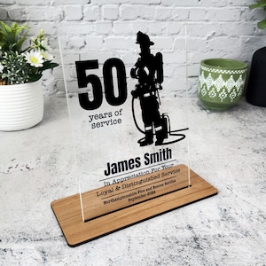 May include: A clear acrylic plaque with a black silhouette of a firefighter standing with a hose. The plaque has the text "50 years of service" and "James Smith" in black. The text "In Appreciation For Your Loyal & Distinguished Service" is also included, along with the text "Northamptonshire Fire and Rescue Service" and "September 2024".