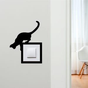 May include: A black cat silhouette climbing on a light switch. The cat is positioned above a white light switch set in a black square frame, mounted on a white wall. The cat's tail is arched, and its body is in a climbing position.