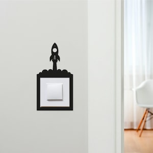 May include: A black rocket-shaped light switch cover is mounted on a white wall. The cover has a square frame around the light switch and a rocket ship design above it. The light switch is white.