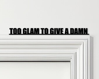May include: Black metal wall art with the phrase "TOO GLAM TO GIVE A DAMN" in a bold, sans-serif font. The text is mounted above a white door frame, creating a striking contrast.