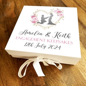 May include: A white keepsake box with a gold heart design and pink floral accents. The box is decorated with a silhouette of a couple getting engaged. The text on the box reads "Amelia & Keith Engagement Keepsakes 12th July 2024".