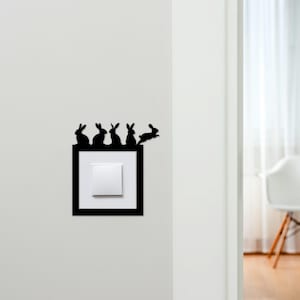 May include: A black decorative light switch cover featuring a silhouette of five rabbits. The rabbits are perched above a white light switch set in a black frame. One rabbit is jumping to the right. The cover is mounted on a white wall.