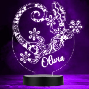 May include: A white, decorative, patterned gecko night light with the name "Olivia" written underneath. The light is on a black base and is illuminated against a purple background.